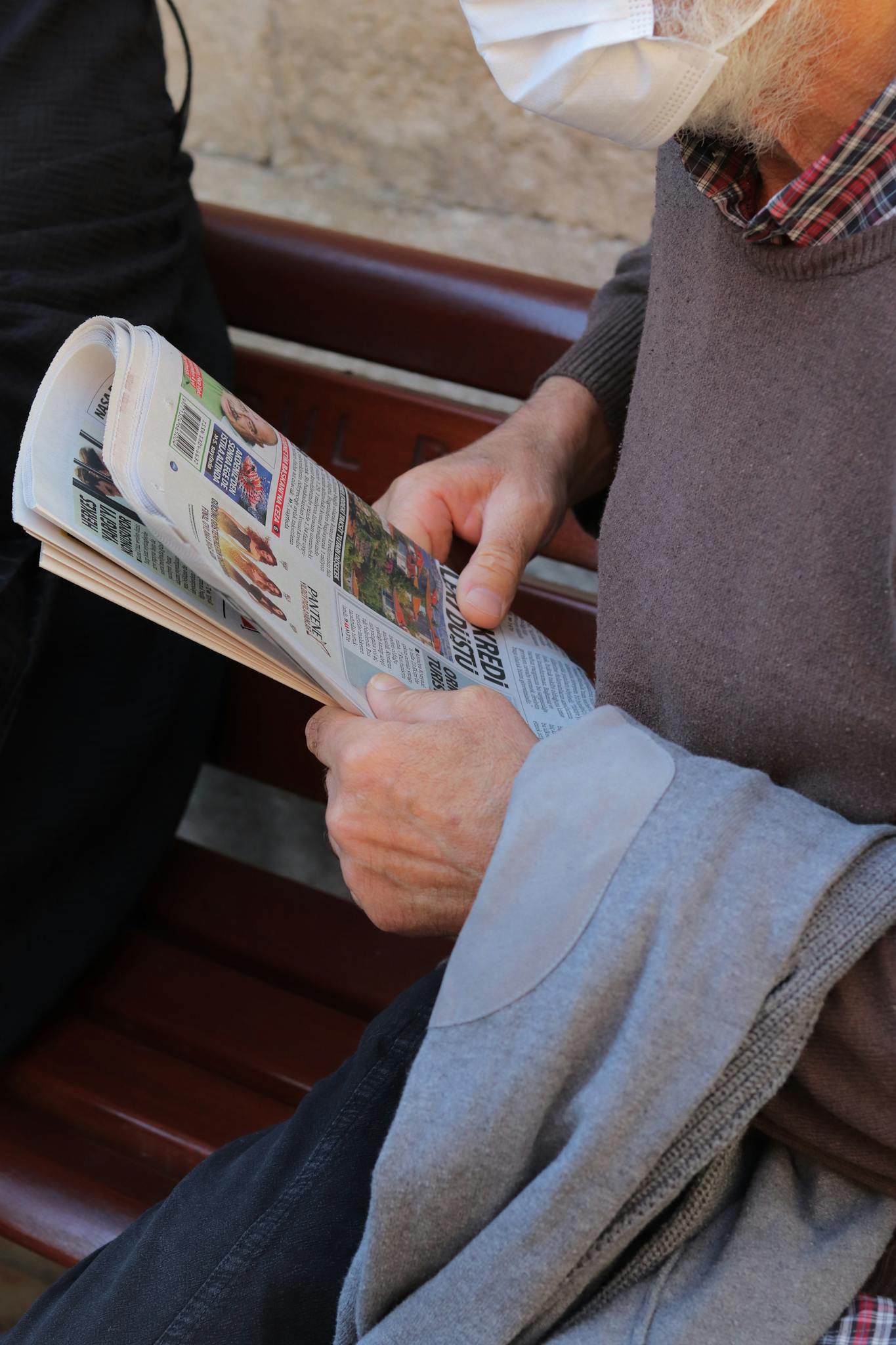 A senior man wearing a mask reads a newspaper on a park bench, focusing intently.