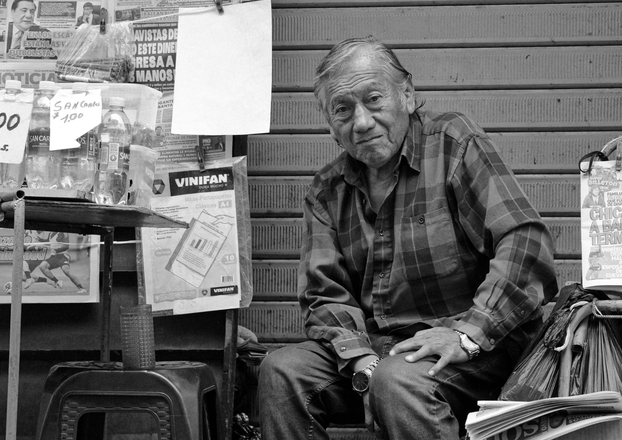 Black and white photo of an elderly man sitting by a street newspaper stand.